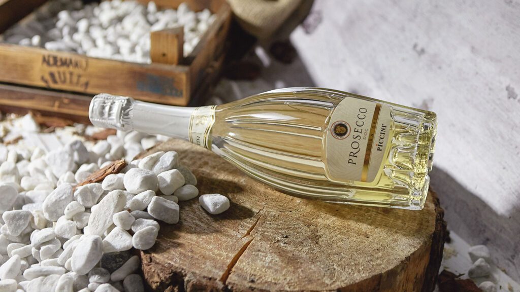 “colombia no es una apuesta, sino una historia que se extiende”, mario piccinni Botella de prosecco doc de piccini recostada sobre un tronco rodeado de piedras blancas decorativas.