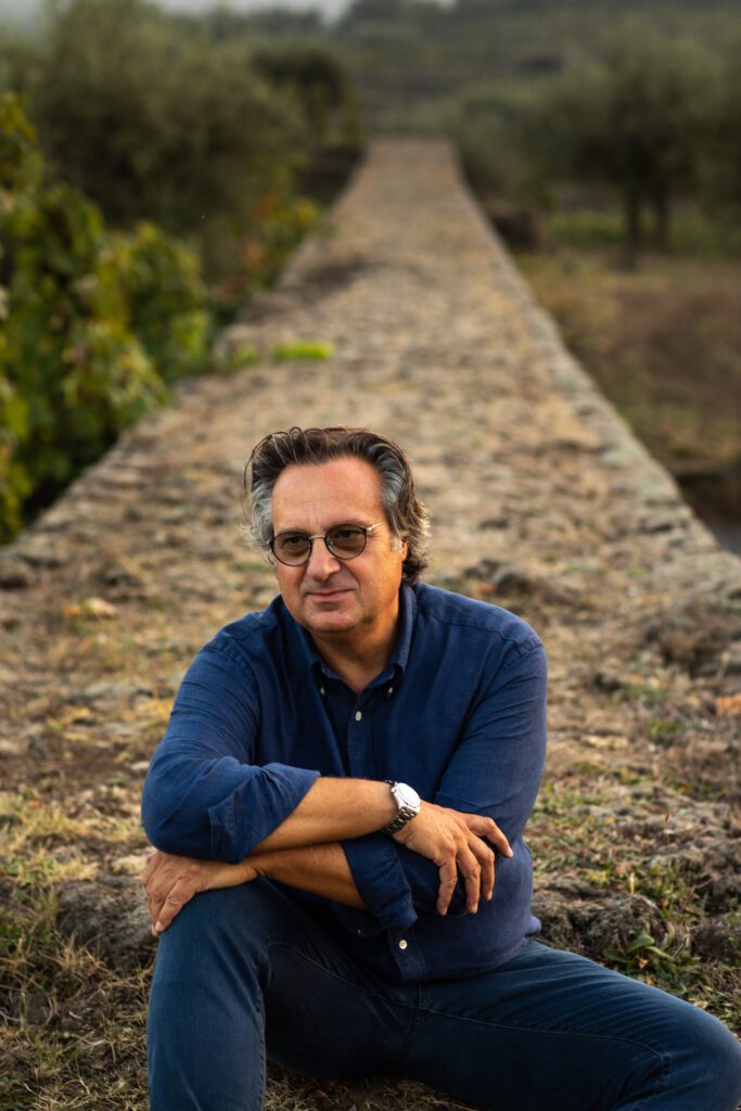 “colombia no es una apuesta, sino una historia que se extiende”, mario piccinni Retrato de mario piccini con camisa azul sentado en un entorno rural al aire libre con viñedos al fondo.