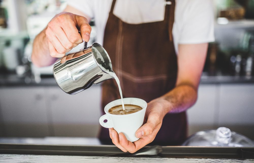 Barista con delantar le pone leche a un capuccino
