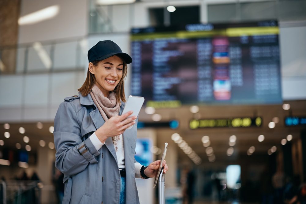 "me lo merezo": por qué cada vez más colombianos usan el viaje como recompensa personal Mensajes de texto de mujer feliz joven en el teléfono celular en el aeropuerto en un viaje.